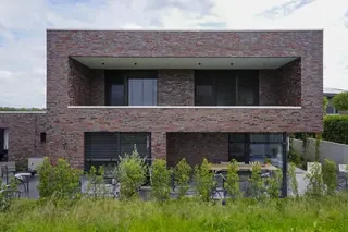 Mischsortierung 198/132 Dünformat Vorderansicht eines modernen, zweigeschossigen Einfamilienhauses mit Flachdach und lebhaft gemischter rot‑braun‑grauer Klinkerfassade. Das Obergeschoss ist zurückgesetzt und bildet eine breite, schattige Loggia mit dunklen Außenjalousien vor großflächigen Fenstern. Davor liegt eine Terrasse mit Sitzbereich, eingelassen hinter einem Streifen junger, niedriger Bepflanzung.
