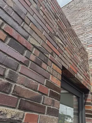 Schräg aufgenommene Detailansicht einer gemischtfarbigen Klinkerwand in Rot‑, Braun‑ und Grautönen. Ein dunkel gerahmtes Fenster ist am rechten Bildrand angeschnitten. Die schmalen Fugen und die lebhafte Farbvariation betonen das handwerkliche Mauerbild.