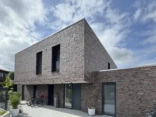 Schräg angeschnittene Vorder‑/Seitenansicht eines modernen zweigeschossigen Flachdachhauses in rot‑grau gemischtem Klinker. Im Erdgeschoss liegt ein zurückgesetzter, überdachter Eingangsbereich; darüber zwei schmale, hohe Fenster mit dunklen Lamellen. Vor dem Haus ein gepflasterter Vorplatz mit abgestellten Fahrrädern und Kübelpflanzen.